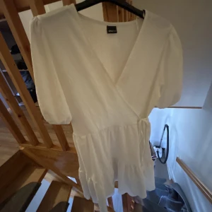 Vit omlottklänning - Säljer en söt vit omlottklänning med korta puffärmar och volangdetaljer. Perfekt för sommarens alla tillfällen. Klänningen har en v-ringad design och är gjord i ett lätt och luftigt material.