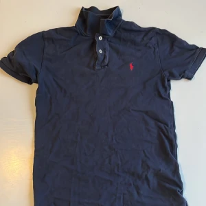 Mörkblå pikétröja från Ralph Lauren - Säljer en klassisk mörkblå pikétröja från Ralph Lauren med korta ärmar och en röd broderad logotyp på bröstet. Tröjan har en knappslå med två knappar och en ribbad krage. Perfekt för en stilren look!