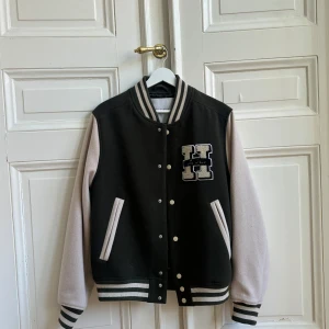 Olivgrön och beige baseballjacka från Les Deux🫒🫒 - Snygg baseballjacka från Les Deux. Jackan har ett stort 'H' broderat på bröstet och 'LES DEUX' på ryggen. Den har ribbade muddar och knappar framtill. Perfekt för en cool och avslappnad stil. Går ner i pris vid snabb affär 