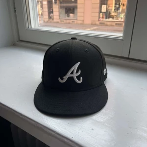 Svart keps från New Era - Snygg svart keps från New Era med en broderad vit logotyp på framsidan. Kepsen har en klassisk design med platt skärm och är en del av 59FIFTY-serien. Perfekt för en stilren look.