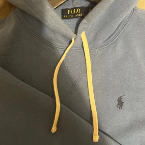 Blå hoodie från Ralph Lauren - Säljer en snygg ljus blå hoodie från Ralph Lauren med vitt snöre. Tröjan ser lite blek ut på bilden och är mer färgglad i verkligheten.