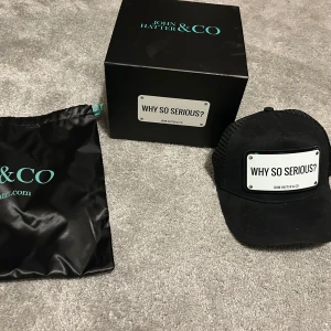 Svart keps från John Hatter & Co - Snygg svart keps från John Hatter & Co med texten 'Why So Serious?' på framsidan. Kepsen har en klassisk design med justerbar passform och nätmaterial på baksidan för extra ventilation. Perfekt för en avslappnad stil.