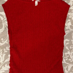  topp från H&M - Säljer en snygg röd topp från H&M. Den har en texturerad yta och är ärmlös, vilket gör den perfekt för varmare dagar. Toppen har en enkel och stilren design som passar till många olika outfits.