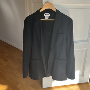 Svart kavaj från Hope - Elegant svart kavaj från Hope med klassisk design. Kavajen har en stilren passform och är perfekt för både formella och avslappnade tillfällen. Unisex. Storlek 40 för kvinnor och 50 för män. 