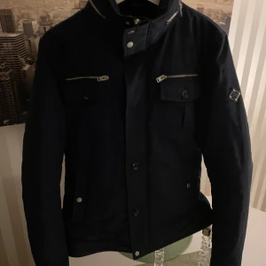 Svart jacka från J.Lindeberg - Snygg blå  jacka från J.Lindeberg med dragkedja och knappar. Jackan har flera fickor med dragkedjor och en stilren design. Perfekt för höst och vår.