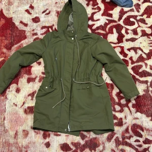Grön parkajacka från Vero Moda - Säljer en snygg grön parkajacka från Vero Moda. Jackan har en justerbar midja med dragsko och en praktisk huva. Perfekt för kyligare dagar med sina långa ärmar och knappar framtill. Ett måste för garderoben!