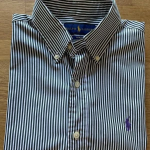  Ralph Lauren skjorta - Snygg randig skjorta från Ralph Lauren i slim fit. Den är vit och grön med en broderad logotyp i lila på bröstet. Perfekta plagget för sommaren.