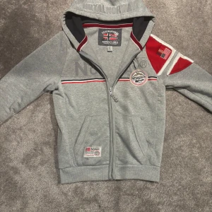 Grå hoodie från Geographical Norway - Säljer en grå hoodie från Geographical Norway med dragkedja och huva. Den har röda och vita detaljer samt en broderad logga på bröstet. Perfekt för kyliga dagar!