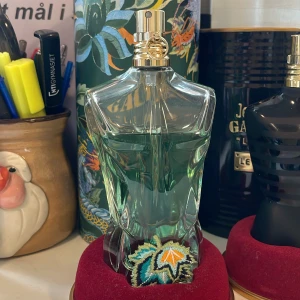Le Beau Parfum från Jean Paul Gaultier - Säljer en elegant Le Beau Parfum från Jean Paul Gaultier. Flaskan är formad som en manlig torso i transparent glas med en grön nyans och har en gyllene kork. Parfymen kommer i en 75 ml flaska och har en tropisk och fräsch doft. Perfekt för den som vill ha något unikt och stilfullt.