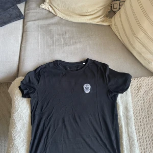 Svart t-shirt med dödskalle från Suspicious Antwerp - Säljer en svart t-shirt från Suspicious Antwerp med en broderad dödskalle på bröstet. T-shirten har en klassisk rund hals och korta ärmar. Perfekt för en cool och avslappnad stil.