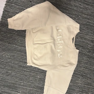 Beige sweatshirt från Bik Bok - Säljer en beige sweatshirt från Bik Bok med texten 'J'adore' broderad på framsidan. Tröjan har långa ärmar och en rund halsringning. Perfekt för en avslappnad stil.