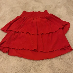 Röd volangkjol från Kappahl - Snygg röd volangkjol från Kappahl med elastisk midja. Perfekt för att ge en färgklick till din outfit. Kjolen har en härlig volangdetalj som ger extra volym och rörelse.