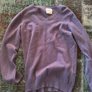 Marinblå kashmirtröja från J.Lindeberg - Säljer en mjuk och stilren lila kashmirtröja från j Lindeberg. Tröjan har en v-ringad design och långa ärmar, perfekt för kyligare dagar. Ribbstickade muddar vid ärmslut och nederkant ger en snygg finish. Passar perfekt för både vardag och mer uppklädda tillfällen.