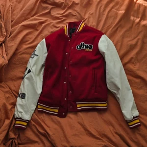 College varsity jacka STWD röd - Snygg röd och vit bomberjacka med broderade detaljer och tryck på ryggen. Jackan har långa ärmar och stängs med knappar. Perfekt för en cool och avslappnad stil.
