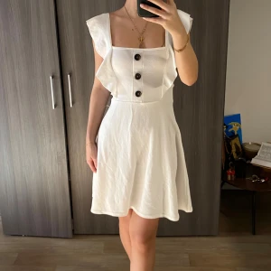 Vit klänning med knappar - Säljer en söt vit klänning med knappar framtill och volangdetaljer vid axlarna. Klänningen har en ärmlös design och en kort längd, perfekt för varma dagar. Den är både stilren och bekväm. Storlek S från boohoo. Endast använd 1 gång.