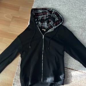 Svart hoodie från Burberry - Säljer en stilren svart hoodie från Burberry med dragkedja och klassiskt rutigt mönster i huvan. Tröjan har en broderad logga på bröstet och långa ärmar. Ej äkta!!!!! 