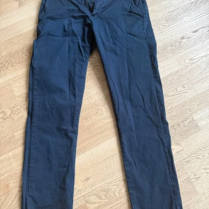 Mörkblå chinos från East West - Säljer ett par mörkblå chinos från East West i regular fit. Byxorna är tillverkade i en bomullsblandning med lite stretch för extra komfort. De har en klassisk design med knappar bak och en stilren look. Perfekta för en snygg och avslappnad stil.