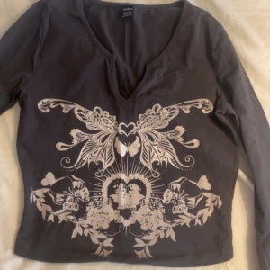 Brun långärmad topp med avcut som magtröja - Säljer en brun långärmad topp med ett dekorativt vitt mönster framtill. Toppen har en v-ringad hals och är perfekt för en stilren look. Passar bra till både jeans och kjol.