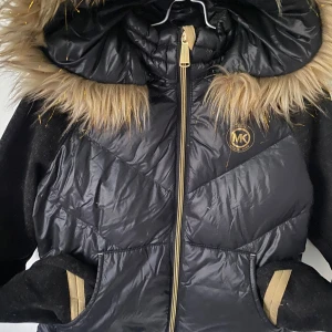 Svart dunjacka från Michael Kors - Snygg svart lättviktsjacka från Michael Kors med en fluffig pälsfodrad huva. Jackan har en dragkedja framtill och är perfekt för kyliga dagar. Den har långa ärmar och en stilren design med diskret logotyp på bröstet. Stl. barn 7/8 år.  Inköpt på Michael Kors-butiken på Macy’s i New York, USA.  Jättefint skick! Använd endast ett fåtal gånger men nu blivit urvuxen.  Hund & katt finns i hemmet