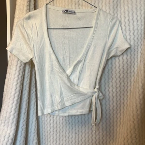 Vit omlottopp från FB Sister - Säljer en stilren vit omlottopp från FB Sister. Toppen har korta ärmar och en knytning i sidan 