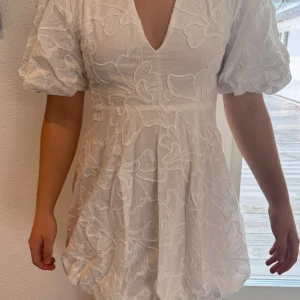 Vit klänning med broderade detaljer - Elegant vit klänning med v-ringad hals och kort puffärm. Klänningen har vackra broderade blommönster och en diskret dragkedja i sidan. Aldrig använd då den är för liten för mig😊   83 cm fr axel ner till kjolens kant.  Midja omkrets ca 73 cm. 