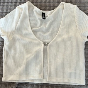 Vit ribbad bolerotopp från Gina Tricot - Säljer en stilren vit ribbad bolerotopp från Gina Tricot. Toppen har korta ärmar och en dekorativ knäppning framtill. Perfekt för att lägga till en touch av elegans till din outfit.