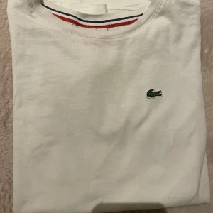 Vit t-shirt från Lacoste - Säljer en vit t-shirt från Lacoste et. T-shirten har ett väldigt litet hål som man ser på en av bilderna men det är inget man tänker på. Jättefin T-shirt verkligen.