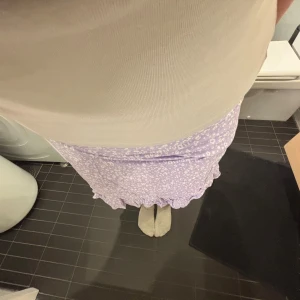 Lila blommig kjol med volang - Söt lila kortkjol med blommigt mönster och volangdetalj längst ner. Perfekt för en somrig look! 🌸säljer pga behov till ny väska så jag samlar pengar💕🏄🏽‍♀️