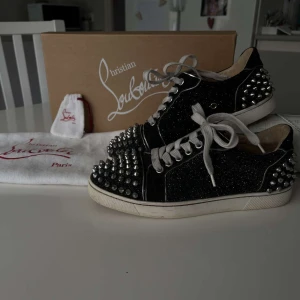 Svarta sneakers med nitar från Christian Louboutin - Snygga svarta sneakers från Christian Louboutin med glittrig yta och nitar på tå och häl. Skorna har vita snören och en vit sula, vilket ger en cool kontrast. Perfekta för att ge din outfit en edgy touch.