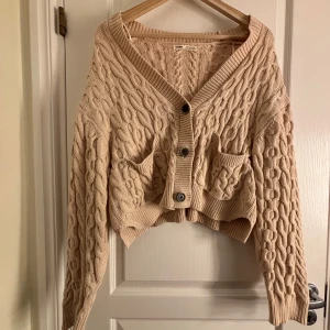 Beige stickad kofta från Pull&Bear - Säljer en snygg beige stickad kofta från Pull&Bear med kabelstickat mönster. Koftan har en croppad passform och knappar framtill. Perfekt för att hålla sig varm och stilren under kyligare dagar.