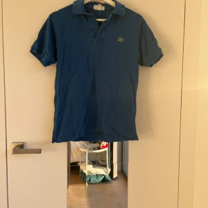Blå pikétröja från Lacoste - Säljer en klassisk blå pikétröja från Lacoste med korta ärmar och den ikoniska krokodilloggan på bröstet. Perfekt för en stilren och avslappnad look. Tröjan har en klassisk passform och är tillverkad i ett bekvämt material.