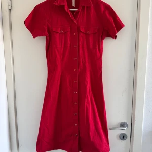 Röd kortärmad klänning från Cubus - Snygg röd klänning från Cubus med korta ärmar och knappar hela vägen ner.  Klänningen har två bröstfickor med knappar och en klassisk krage. (Katt o kanin finns i hemmet) 
