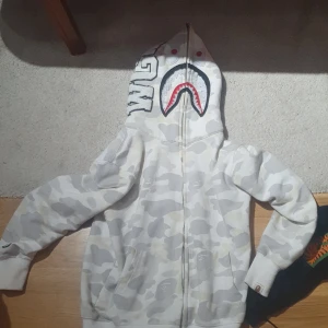 Vit camo hoodie från A Bathing Ape - Säljer en vit camo hoodie från A Bathing Ape med det ikoniska hajmotivet på huvan. Hoodien har dragkedja och är perfekt för en streetwear-look. Den är i storlek M och har en unik design med broderade detaljer.