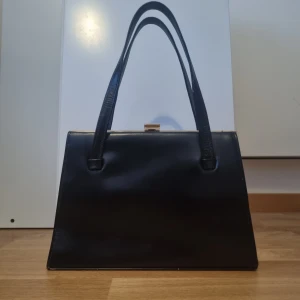 Svart handväska i skinn - Elegant svart handväska i skinn med en stilren design. Väskan har en metallram och ett praktiskt innerfack med dragkedja. Perfekt för att bära dina nödvändigheter med stil.