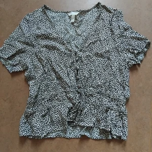 Beige Blus med knappar - En kortärmad blus i ljusbeige med svart prickigt mönster. Knappar framtill och markerad midja med resår och dragsko som man kan justera. Från H&M, storlek 40 men sitter som 36/38. Toppskick!