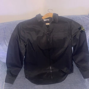 Svart overshirt från Stone Island - Svart overshirt från Stone Island med klassisk krage, dragkedja framtill och bröstficka. Ikonisk logga på vänster ärm. Snygg och clean design i polyester, perfekt för lager-på-lager. Passar dig som gillar streetwear och stilrena plagg.  Pris kan diskuteras!!