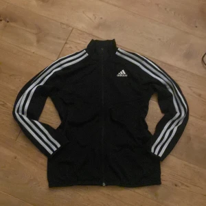 Svart Adidas track jacket med vita ränder - Svart Adidas track jacket med klassiska vita ränder längs ärmarna och Adidas-logga på bröstet. Jackan har dragkedja framtill, hög krage och är tillverkad i ett mjukt, sportigt material. Perfekt för en sportig och chill stil.