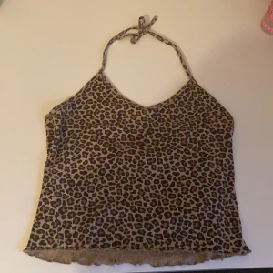 Halterneck leopard topp - Halterneck leopardmönstrad topp från H&M med knyt i nacken. Toppen är beige och brun med svarta detaljer, har v-ringning och volangkant nertill. Perfekt för dig som vill sticka ut med ett djurmönster och gillar trendiga detaljer.