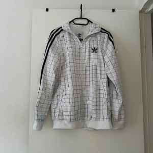 Vit rutig Adidas vindjacka - Vit vindjacka från Adidas med svart rutmönster och klassiska svarta ränder på axlarna. Jackan har huva, dragkedja framtill och två fickor. Har blå fläckar från tvätt. 