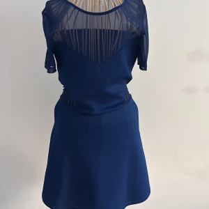 Mörkblå kortklänning med meshdetaljer - Supersnygg mörkblå kortklänning med transparent mesh upptill, i ärmarna och i sidorna vid midjan. Klänningen har korta ärmar och en lätt utställd kjol. Materialet känns mjukt och stretchigt, perfekt för dig som vill sticka ut lite extra. Stl 36