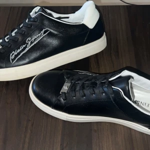 Svarta Plein Sport sneakers i skinn - Snygga svarta sneakers från Plein Sport med vit sula och vit häl. Skorna har snörning och märkets logga broderad på sidan. Tillverkade i glansigt skinn med rund tå och klassisk låg profil. Perfekta för dig som gillar stilrena och sportiga skor.