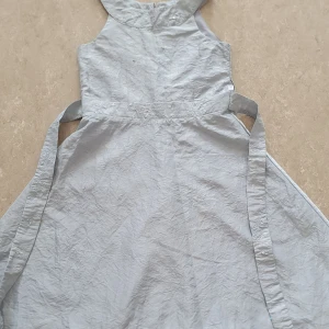 Klänning - Supersöt grå klänning från Lindex med broderade detaljer och små paljetter i midjan. Klänningen har knytband i sidorna och dragkedja bak. Klänningen har en liten fläck på och bandet bakom klänningen har gått av. Perfekt för dig som gillar en enkel men ändå glittrig look.