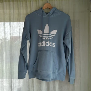 Ljusblå  Adidas hoodie - Snygg ljusblå hoodie från Adidas med stor vit Trefoil-logga framtill. Tröjan har huva med snörning och en klassisk känguruficka. Tillverkad i mjuk bomullsmix och har långa ärmar med ribbade muddar. Perfekt för en chill och sportig look. Mkt bra skick!