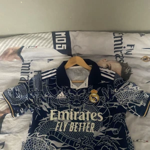 Real Madrid x Adidas blå draktröja S - Säljer en fet Real Madrid fotbollströja från Adidas i marinblått med unikt drakmönster över hela tröjan. Klassisk krage, vita detaljer och klubbmärke på bröstet. Materialet är lätt och andas, perfekt för match eller träning. Emirates Fly Better tryck på framsidan.  Skriv för att diskutera pris !