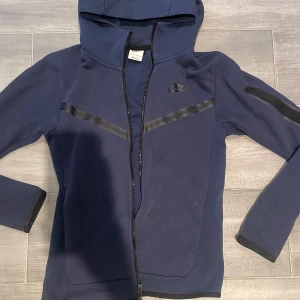 Blå nike tech jacka - Snygg blå hoodiejacka från Nike i storlek XS. Jackan har hel dragkedja, huva och svarta detaljer över bröstet samt en ficka med dragkedja på ärmen. Använd ett fåtal gånger men köpt på Plick! 