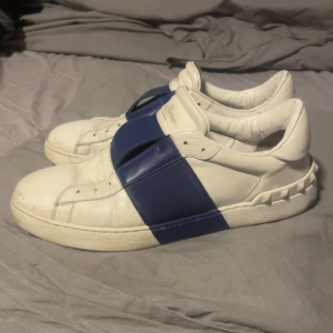Valentino Open sneakers vit/blå - Snygga Valentino Open sneakers i vitt skinn med bred blå rem över vristen. Klassisk rund tå och platt sula med ikoniska nitar baktill. Perfekta för dig som gillar stilrena men ändå unika skor.