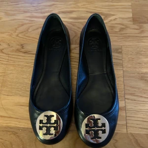 Nya Svarta ballerinaskor från Tory Burch - Stilrena svarta ballerinaskor från Tory Burch i skinn med rund tå och ikonisk guldfärgad logga framtill. Perfekta för dig som gillar klassisk och elegant stil med en touch av lyx.