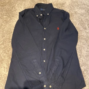 Mörkblå skjorta från Ralph Lauren - Snygg mörkblå skjorta från Ralph Lauren i slim fit-modell. Skjortan har klassisk button down-krage, vita knappar och den ikoniska röda broderade loggan på bröstet. Tillverkad i bomull och med långa ärmar. Perfekt för en clean och stilren look. Aldrig tvättad för inte behövts använd 8 gånger