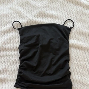 Svart bandeau tubtopp med rynk - Säljer en svart bandeau tubtopp med tunna axelband och snygga rynkade detaljer på sidorna. Toppen är figurnära och har en enkel, stilren look som passar perfekt till jeans eller kjol. Gjord i mjukt material för en bekväm känsla.
