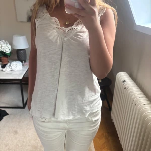 Vit spetsdetaljerad linnetopp - Supersöt vit linnetopp med broderade spetsdetaljer längs v-ringningen och axlarna. Toppen är ärmlös och har en avslappnad passform, perfekt för varma dagar. Materialet känns mjukt och luftigt, och spetsen ger en romantisk touch.
